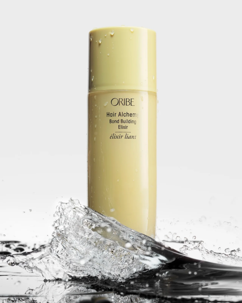 Pale yellow bottle of Oribe Hair Alchemy Bond Building Elixir surrounded by splashing water.