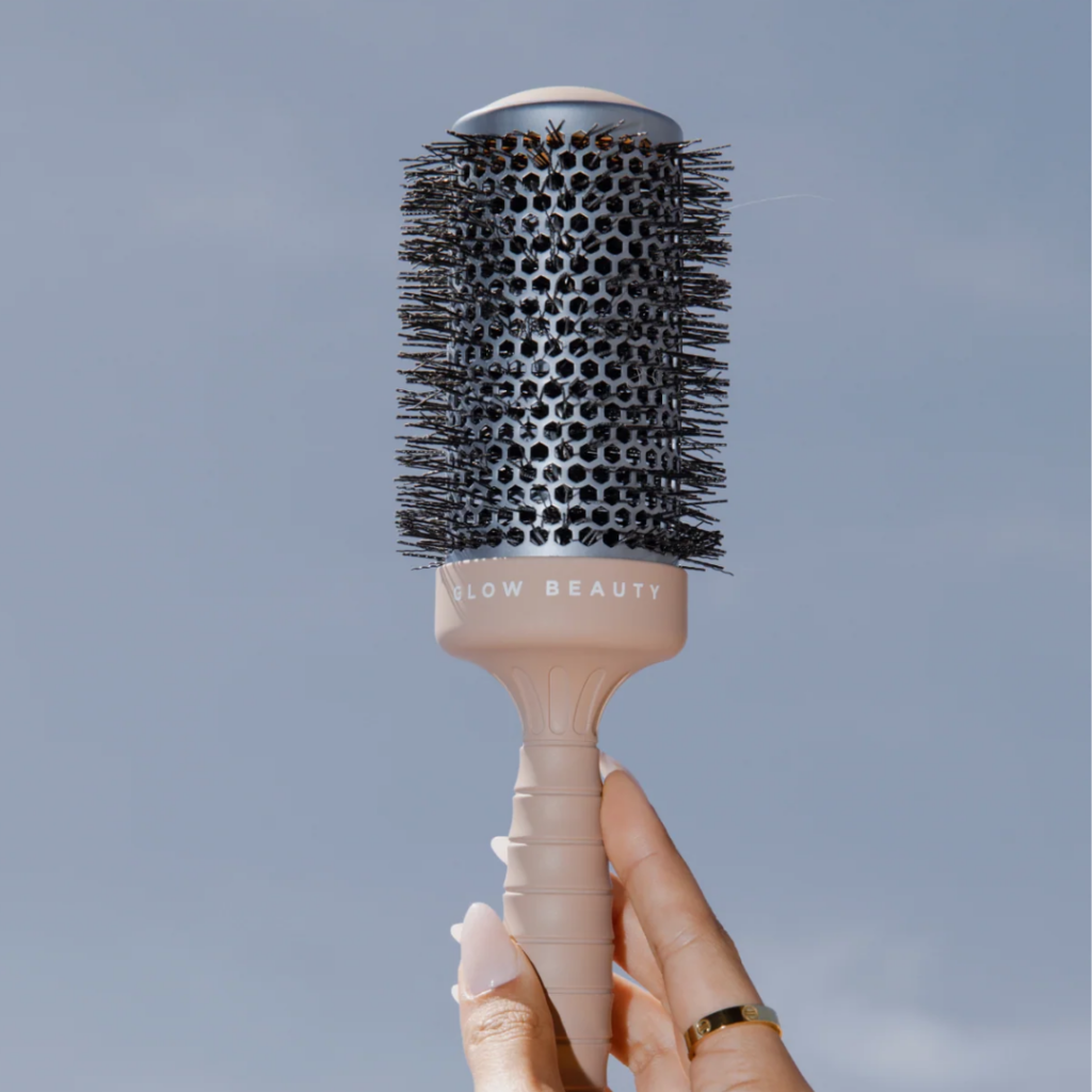 Hand holding the Glow Beauty round styling brush against a soft neutral background for an October 2026 product spotlight.